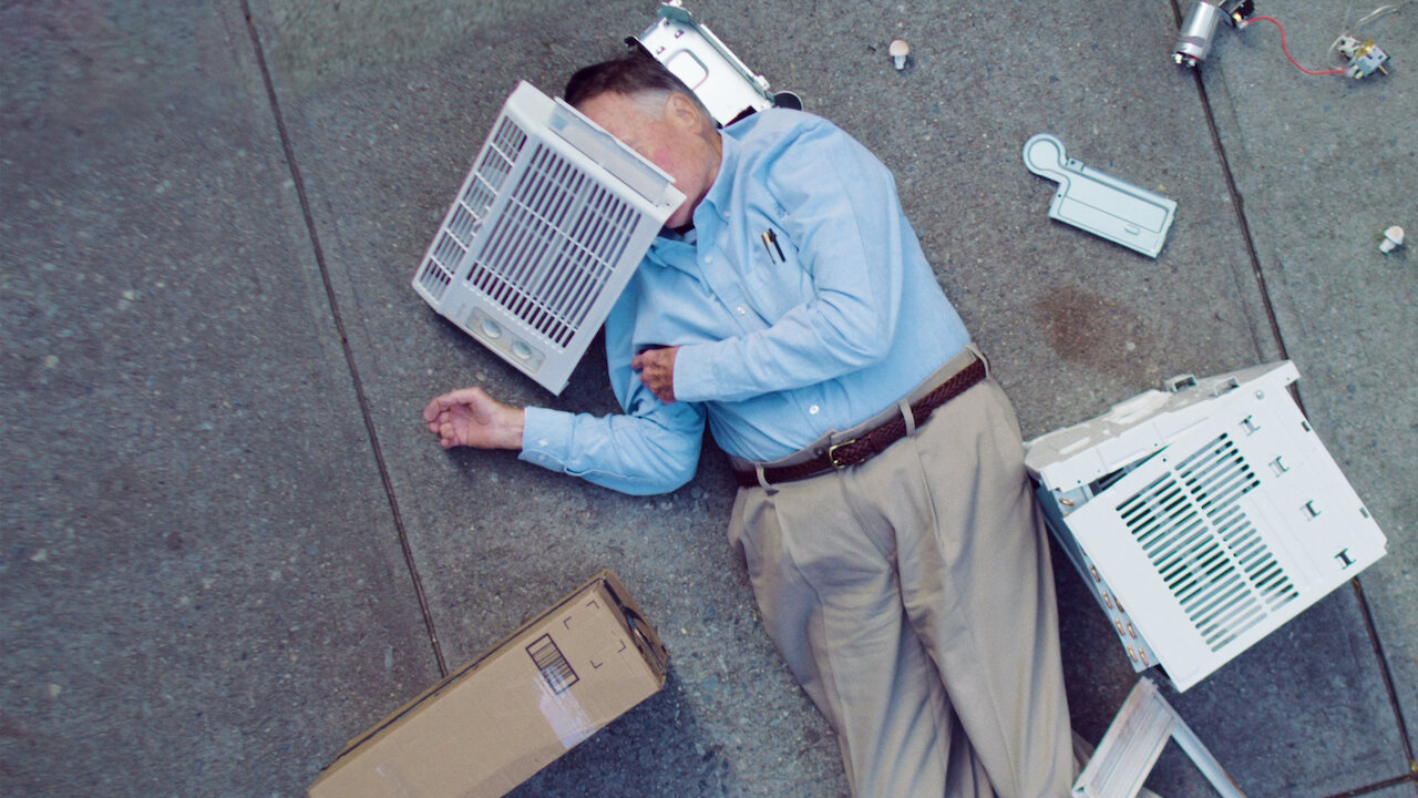 Image is from the film 'Dick Johnson is Dead' (2020). An older man dressed in a polo and khakis lies on the ground outside, a box at his side and the pieces of a broken desktop computer surrounding him. Two of the computer pieces lay on either side of his head, one obscuring his face.